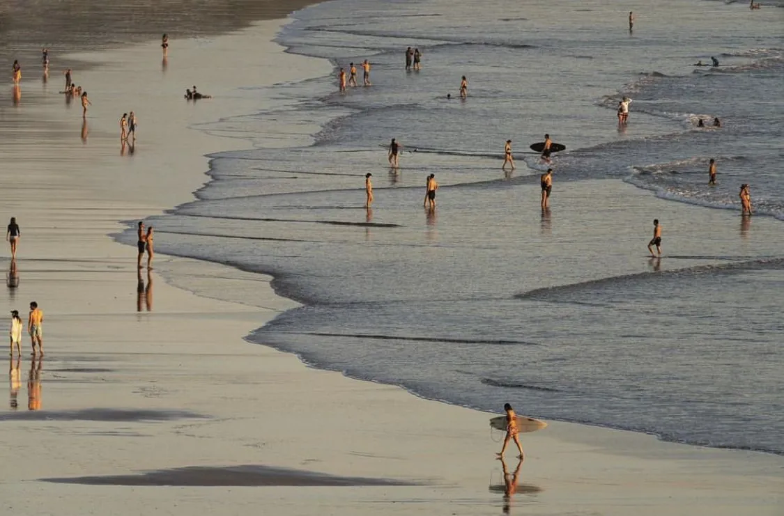 Golden-hour beach with swimmers and a surfer walking along the shoreline